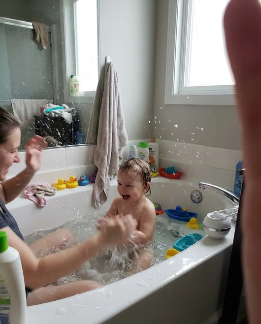Child in a bathtub with toys and a woman sitting beside them, with a PureToddle Faucet Filter on the edge of the bath.